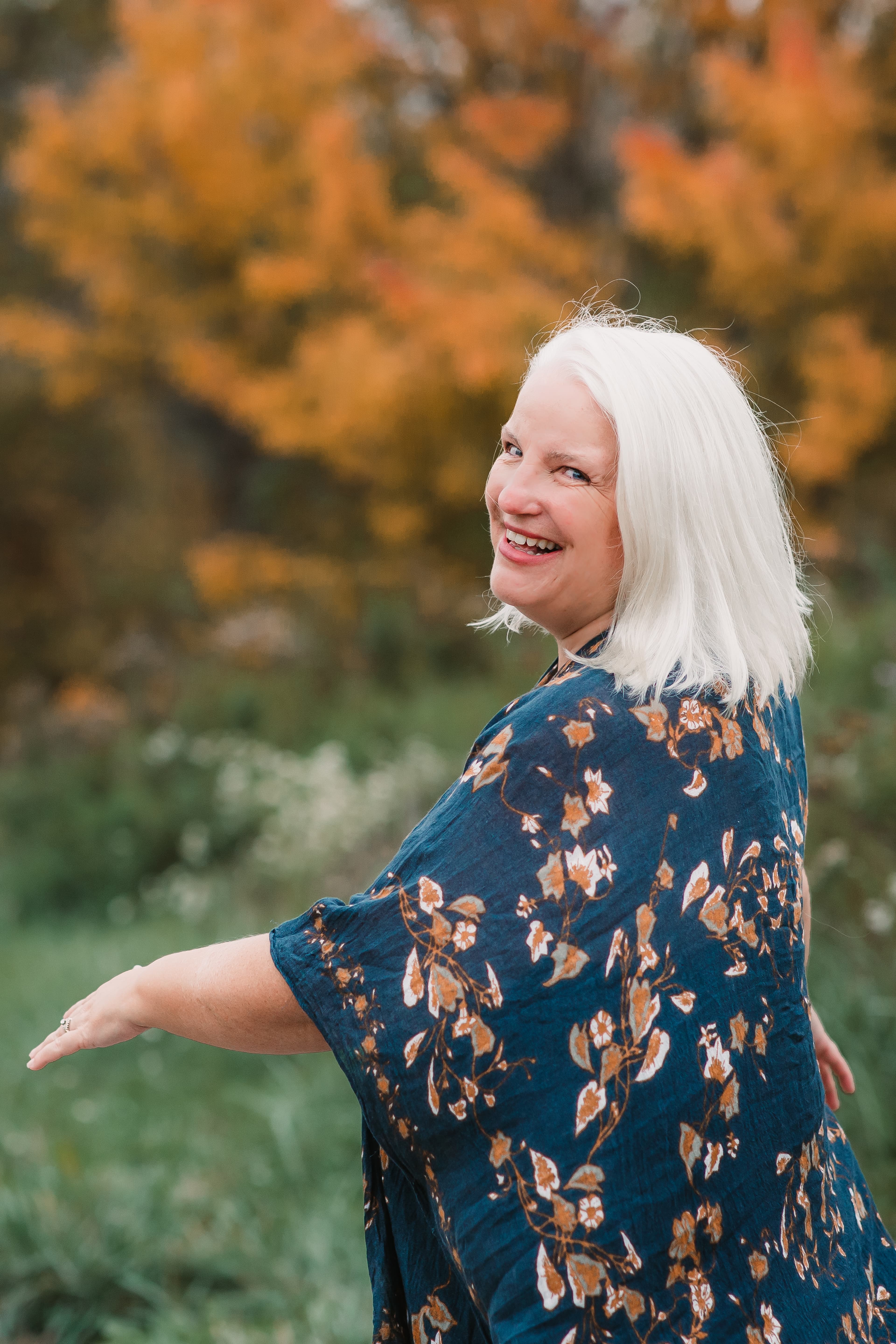 Nola Simon looking over her shoulder with a warm smile, wearing a teal floral kimono against autumn foliage